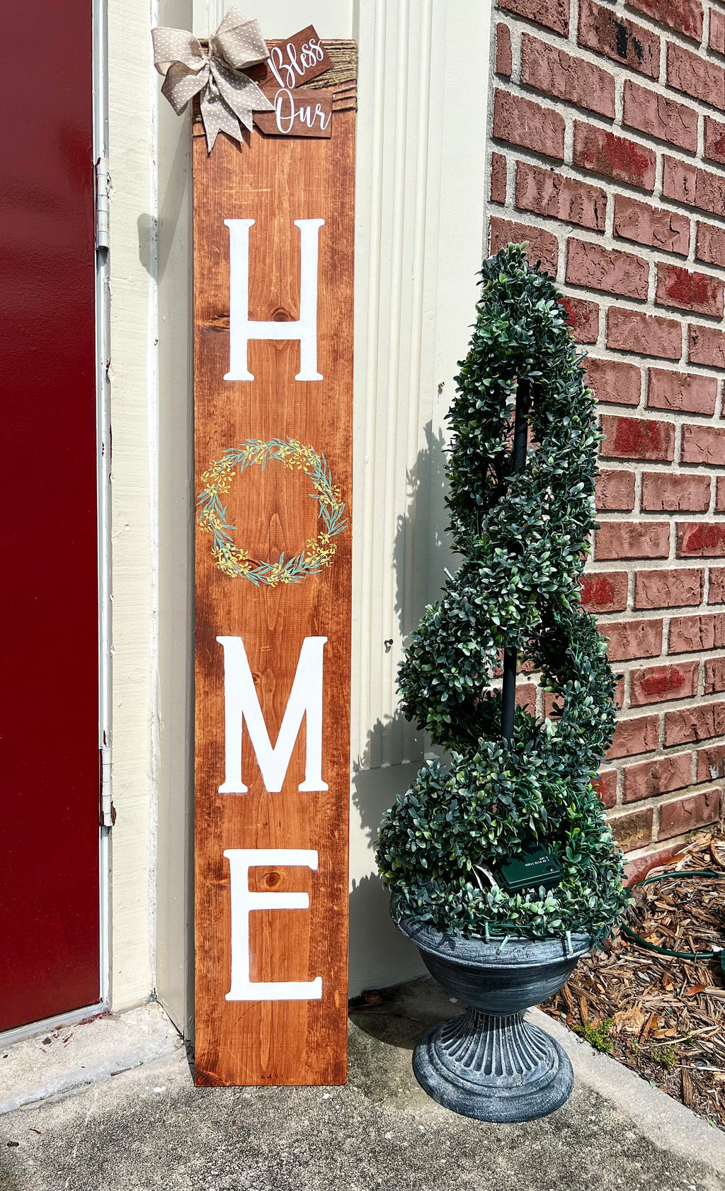 Bless Our Home porch leaner sign in light walnut stain full view with boxwood topiary and brick background
