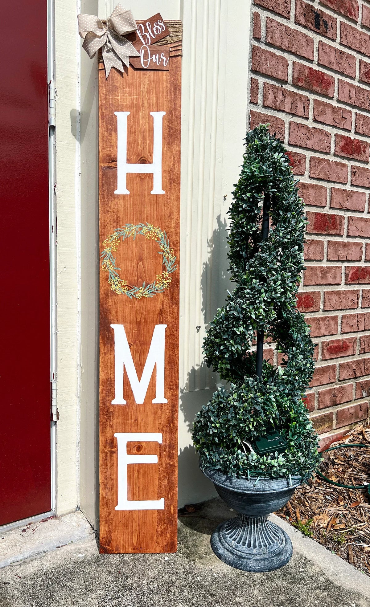 Bless Our Home porch leaner sign in light walnut stain full view with boxwood topiary and brick background