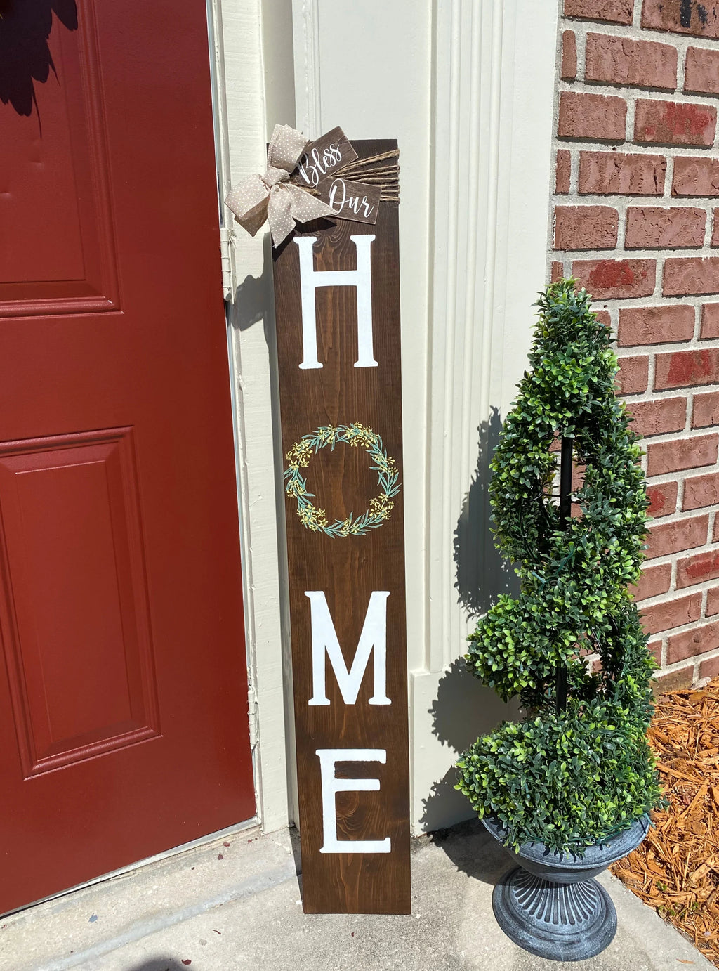 Bless Our Home porch leaner sign in dark walnut with burlap bow against red door full length view