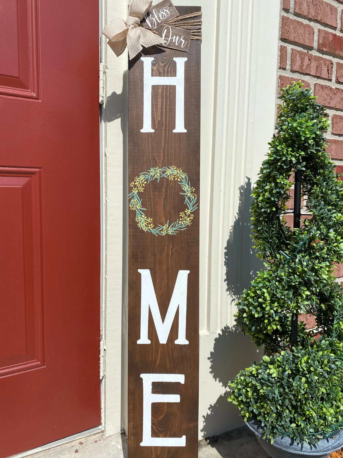 Bless Our Home porch leaner sign in dark walnut leaning against brick wall with boxwood topiary