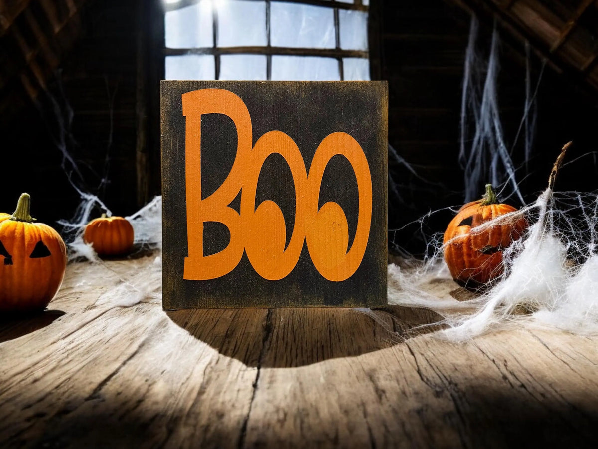 Boo black wood block sign with orange lettering on dark wood table flanked by glowing jack-o-lanterns in moody Halloween setting