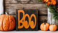 Boo black wood block sign with orange lettering on rustic wood porch shelf with large orange pumpkins and fall foliage