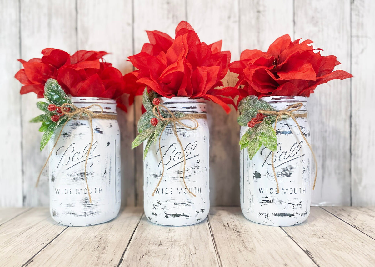 Three distressed white farmhouse mason jars with red poinsettias and holly close up