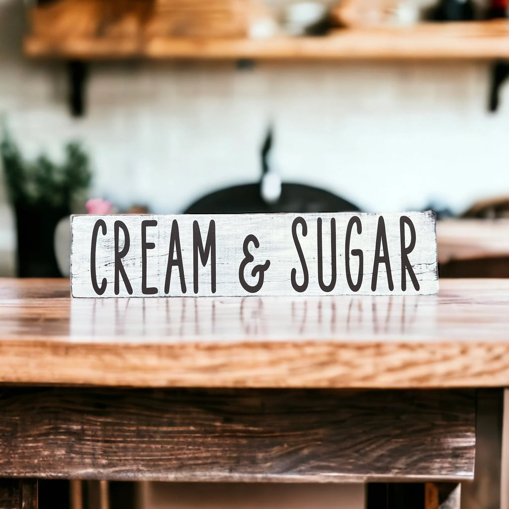 Cream and Sugar distressed white farmhouse wood sign on wood kitchen counter with wicker basket and black canisters in background