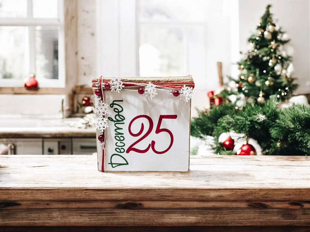December 25 Christmas wood block sign with red 25 and green December script jute top and snowflake bead garland on rustic wood surface with red ornament and pine sprig close up