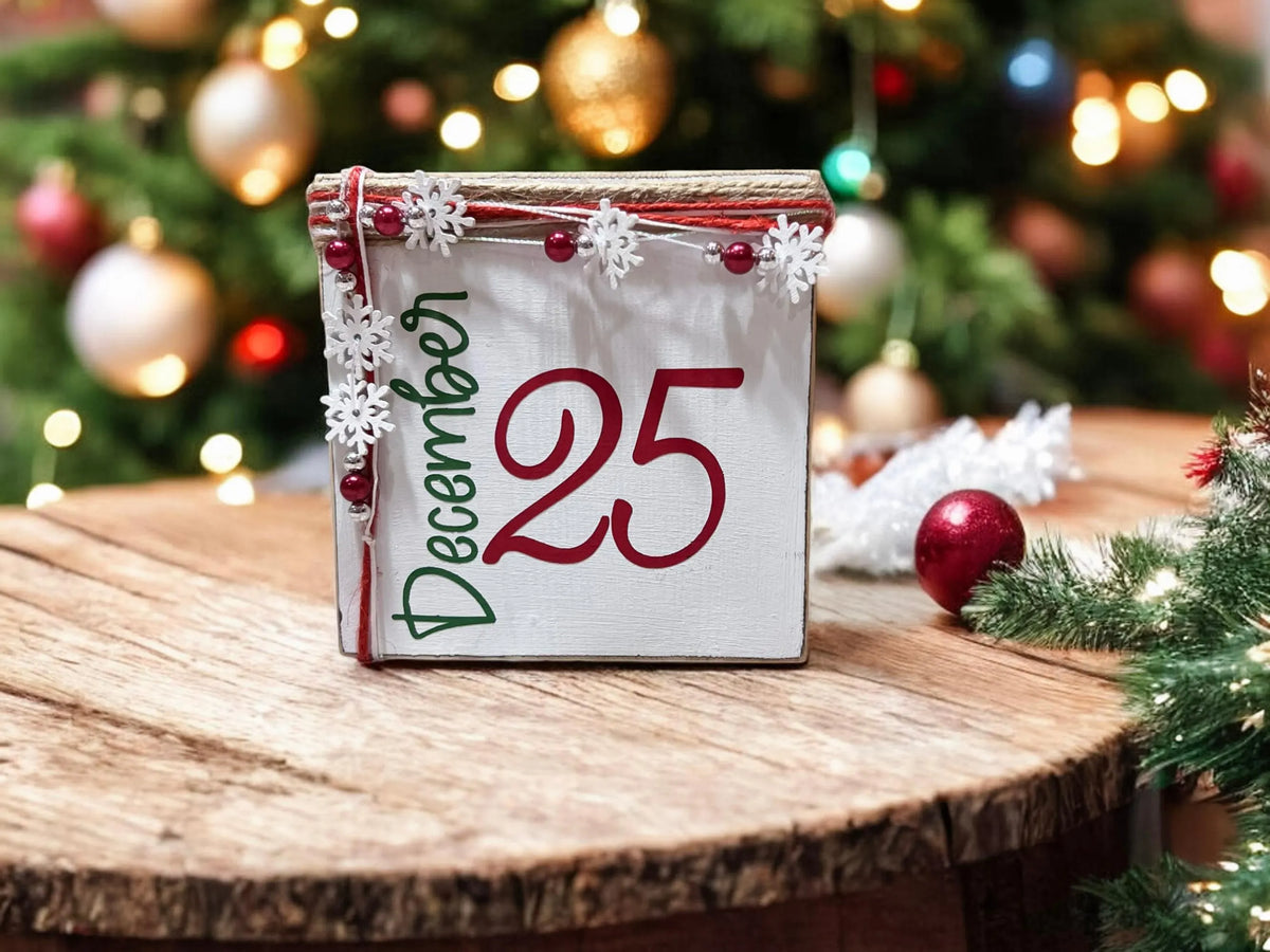 December 25 Christmas wood block sign with red 25 and green December script jute top and snowflake bead garland on white wood surface with lit Christmas tree in farmhouse kitchen background