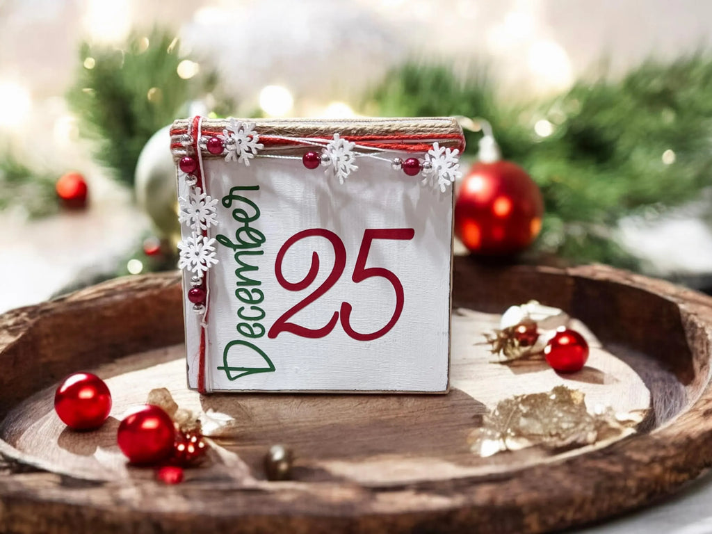 December 25 Christmas wood block sign with red 25 and green December script jute top and snowflake bead garland on rustic wood kitchen counter with lit Christmas tree and pine cones