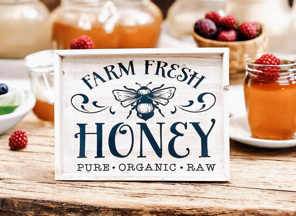 Farm Fresh Honey framed reclaimed wood sign with navy bee graphic and chalk paste lettering in distressed whitewash frame on rustic wood surface with small honey jar and blueberry close up