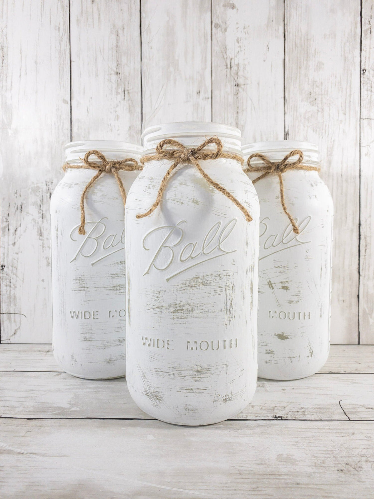 Set of 3 distressed white Ball mason jars with jute twine bows wide view on white wood surface