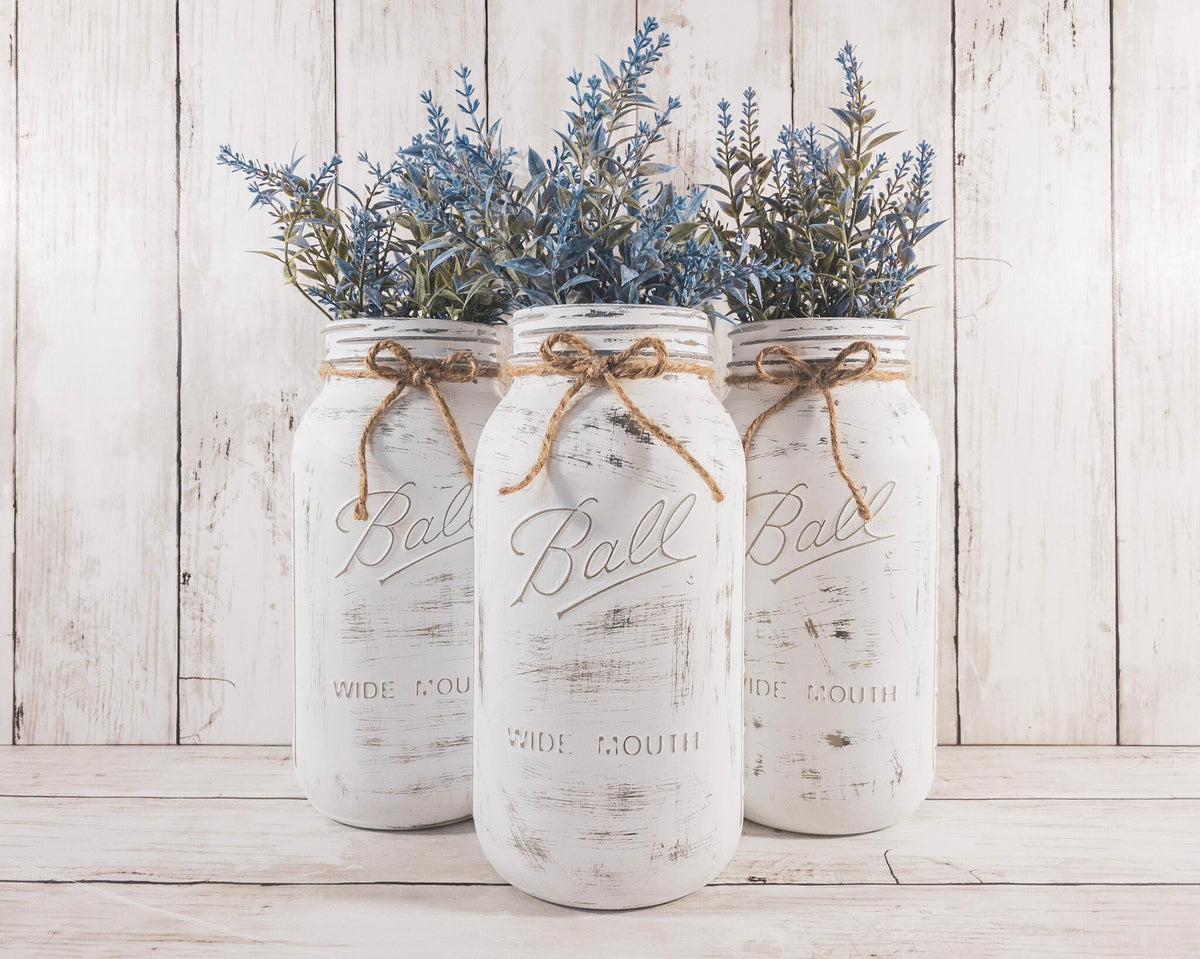 Set of 3 distressed white Ball mason jars with jute twine bows and blue lavender greenery on white wood surface