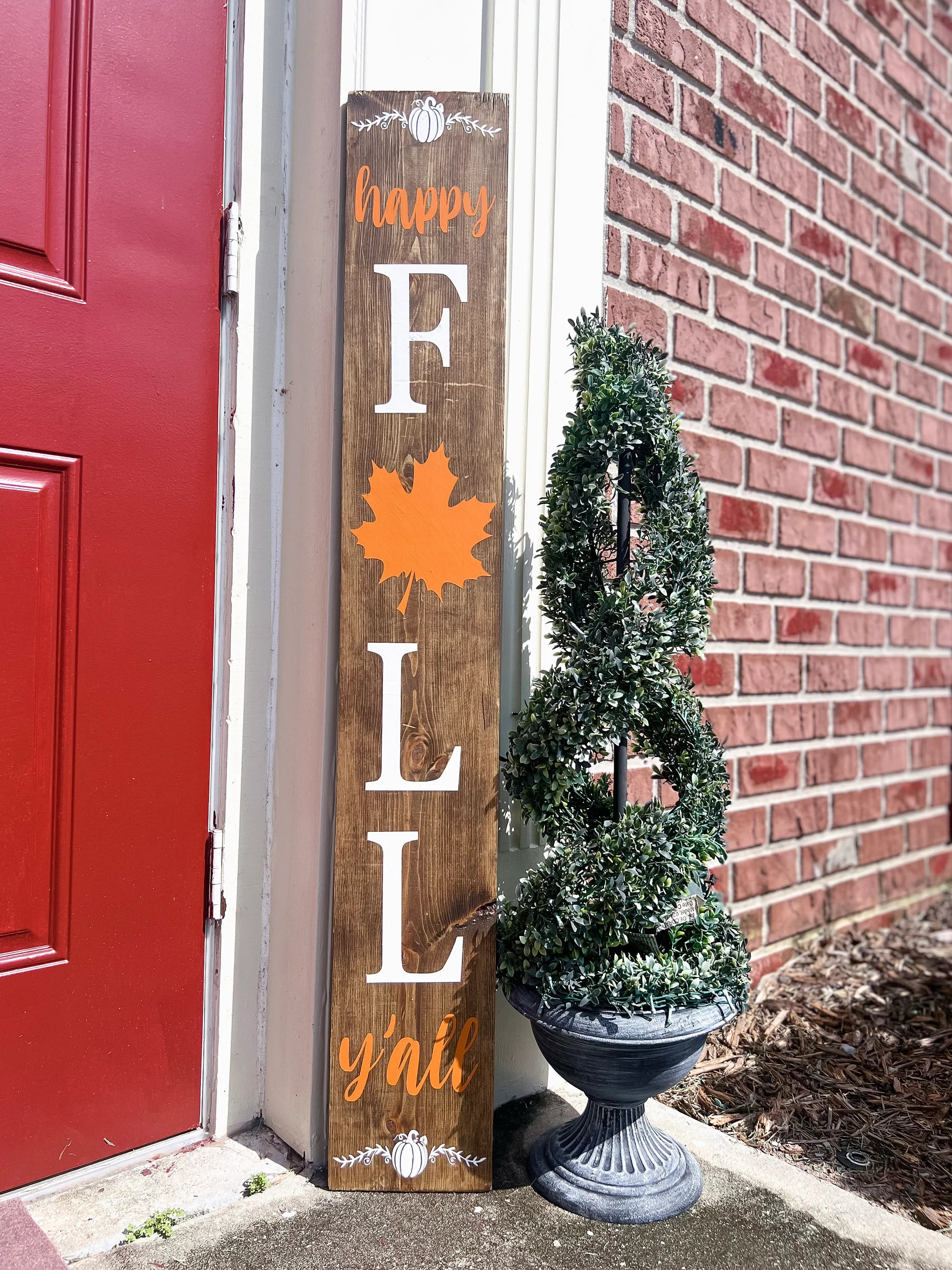 Happy Fall Yall dark walnut porch leaner sign with white serif FALL letters orange maple leaf and orange script leaning against white door frame beside spiral boxwood topiary on brick porch full length view