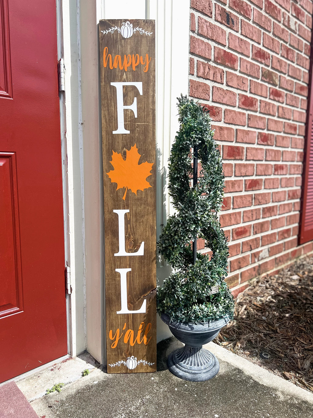 Happy Fall Yall dark walnut porch leaner sign with white serif FALL letters orange maple leaf and orange script leaning against white door frame beside spiral boxwood topiary on brick porch side angle