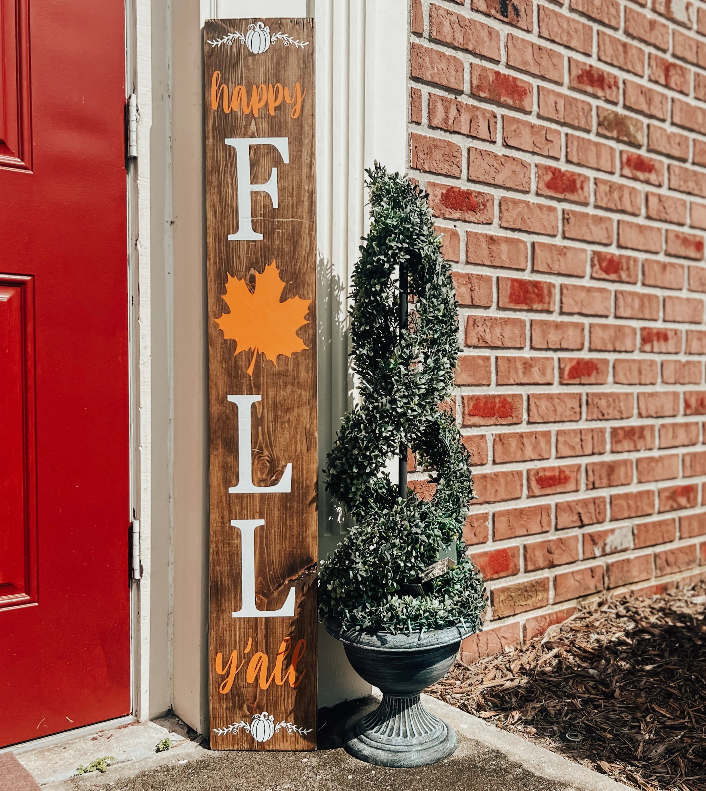 Happy Fall Yall dark walnut porch leaner sign with white serif FALL letters orange maple leaf and orange script leaning against white door frame beside spiral boxwood topiary on brick porch angled view