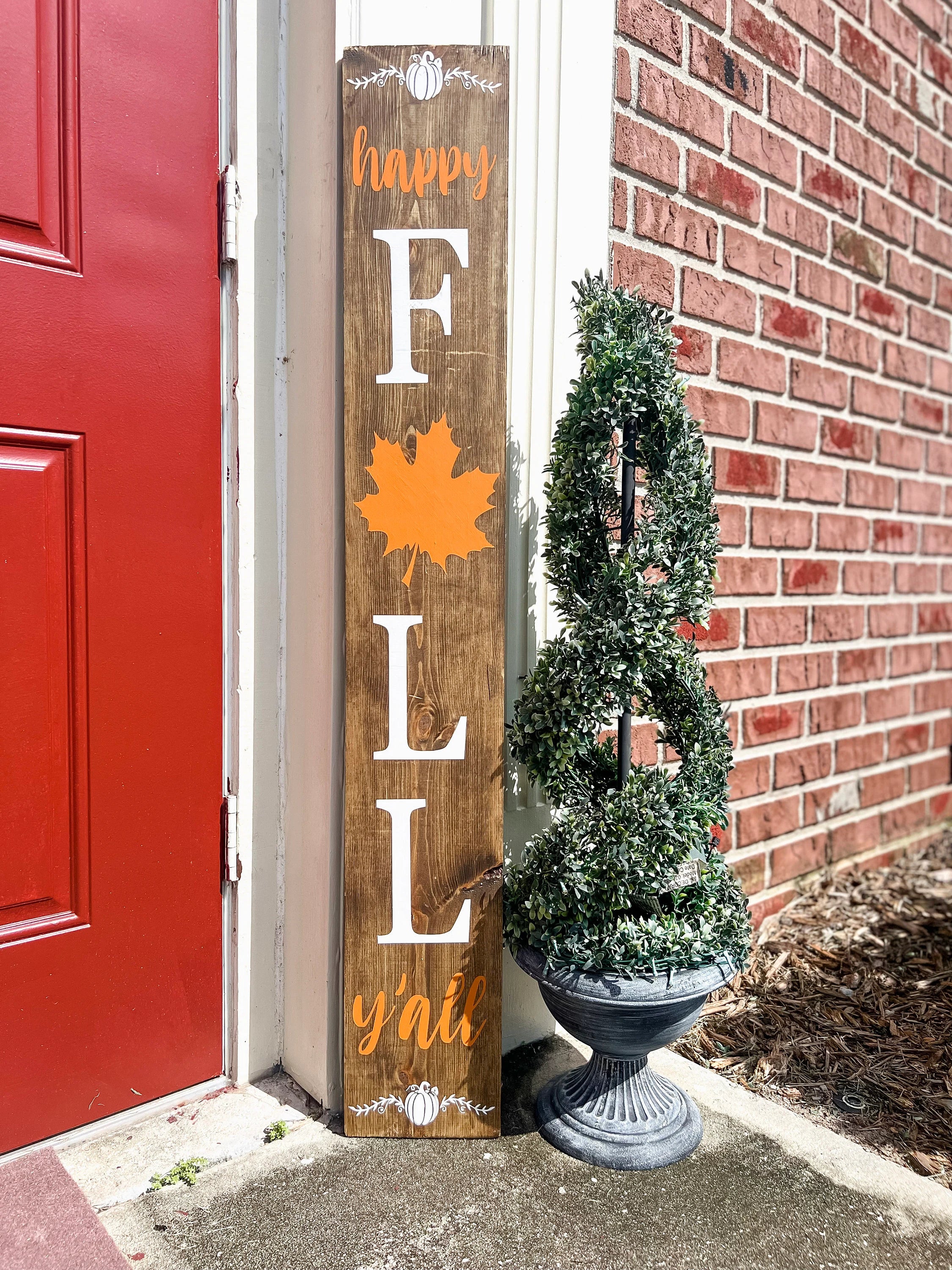 Happy Fall Yall dark walnut porch leaner sign with white serif FALL letters orange maple leaf and orange script leaning against white door frame beside spiral boxwood topiary on brick porch wide shot