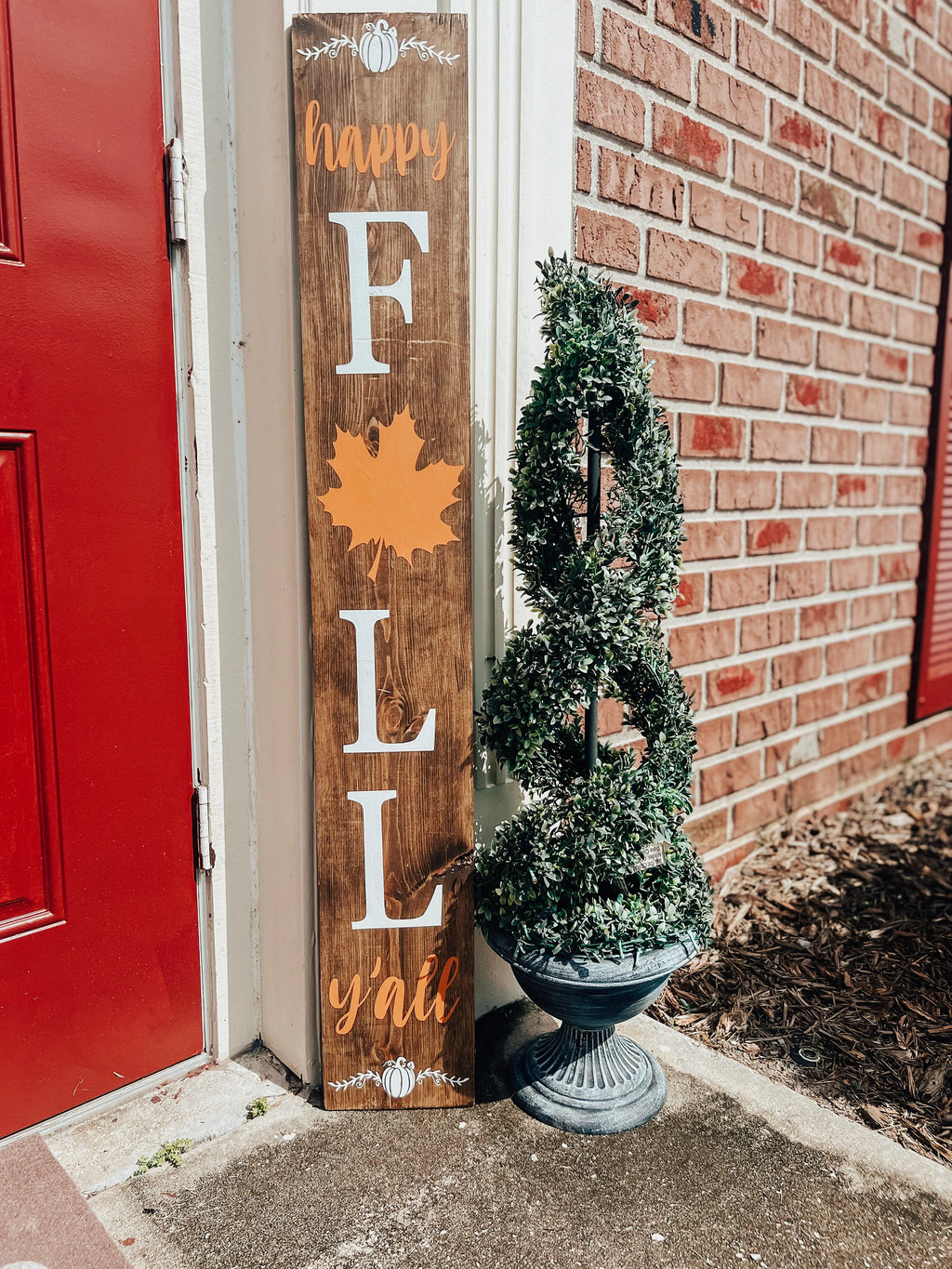 Happy Fall Yall dark walnut porch leaner sign with white serif FALL letters orange maple leaf and orange script leaning against white door frame beside spiral boxwood topiary on brick porch hero shot