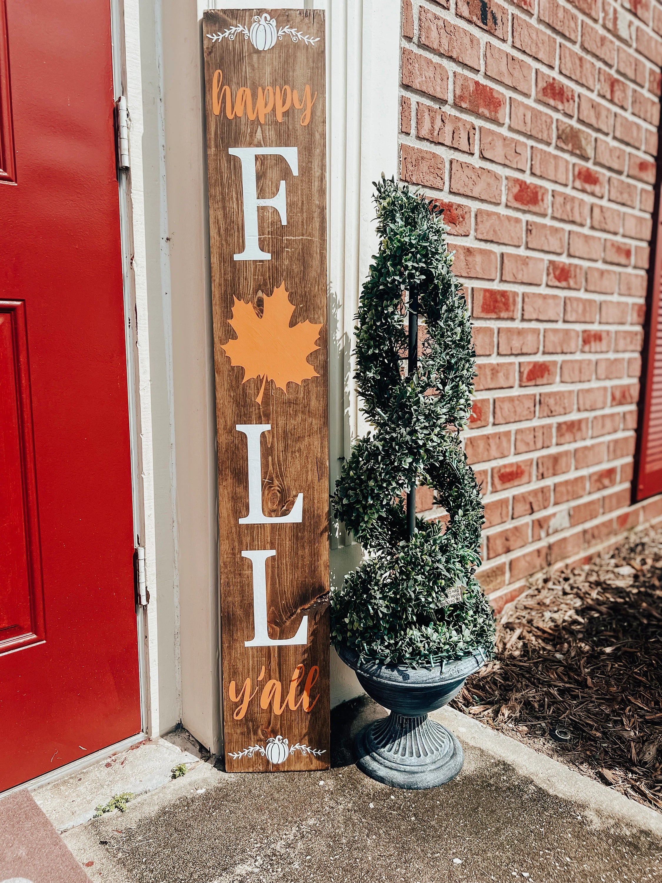 Happy Fall Yall dark walnut porch leaner sign with white serif FALL letters orange maple leaf and orange script leaning against white door frame beside spiral boxwood topiary on brick porch hero shot