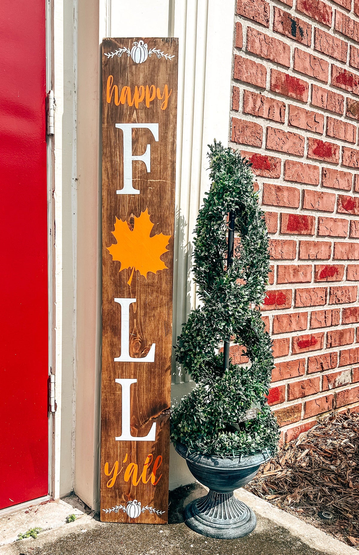 Happy Fall Yall dark walnut porch leaner sign with white serif FALL letters orange maple leaf and orange script leaning against white door frame beside spiral boxwood topiary on brick porch lifestyle shot