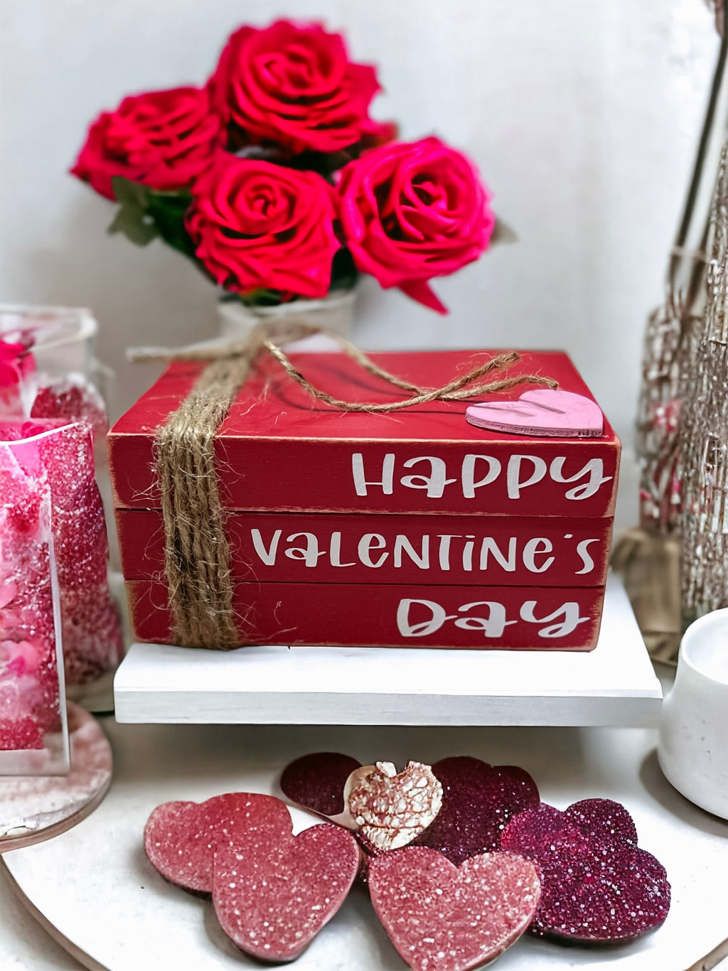 Happy Valentine's Day red wood book stack with jute twine and pink heart on white tray with pink carnations and candy jars