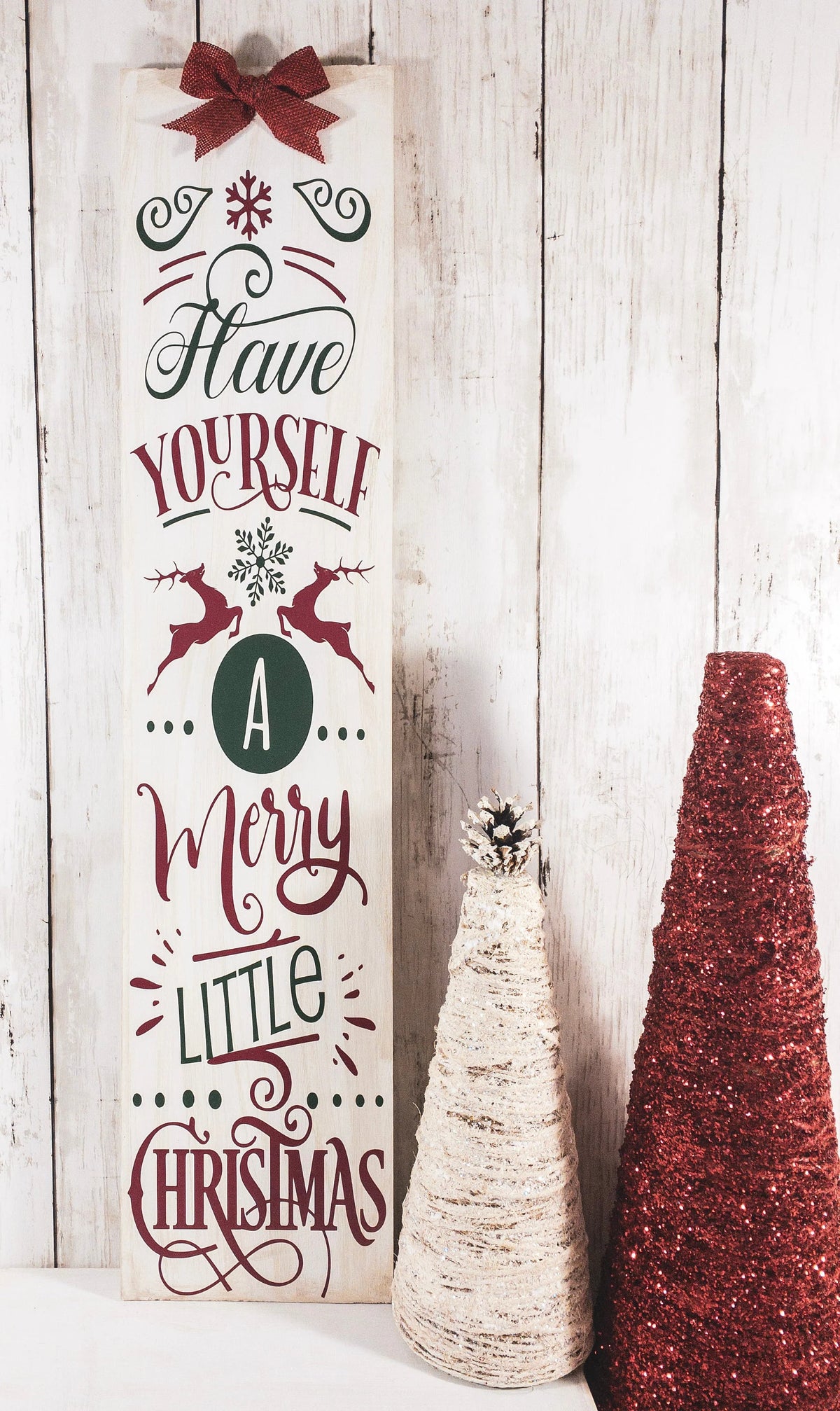 Have Yourself a Merry Little Christmas rustic wood porch leaner sign with red bow leaning against white door beside boxwood topiary wide view