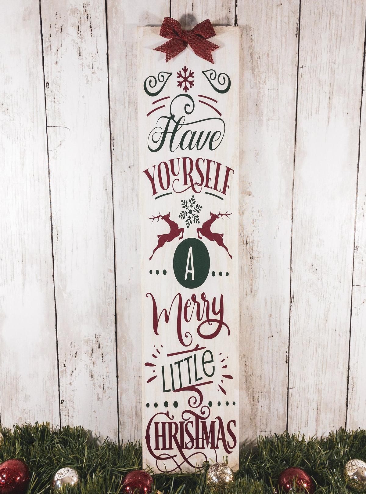 Have Yourself a Merry Little Christmas rustic wood porch leaner sign with burlap bow on white wood wall beside glitter Christmas trees wide view