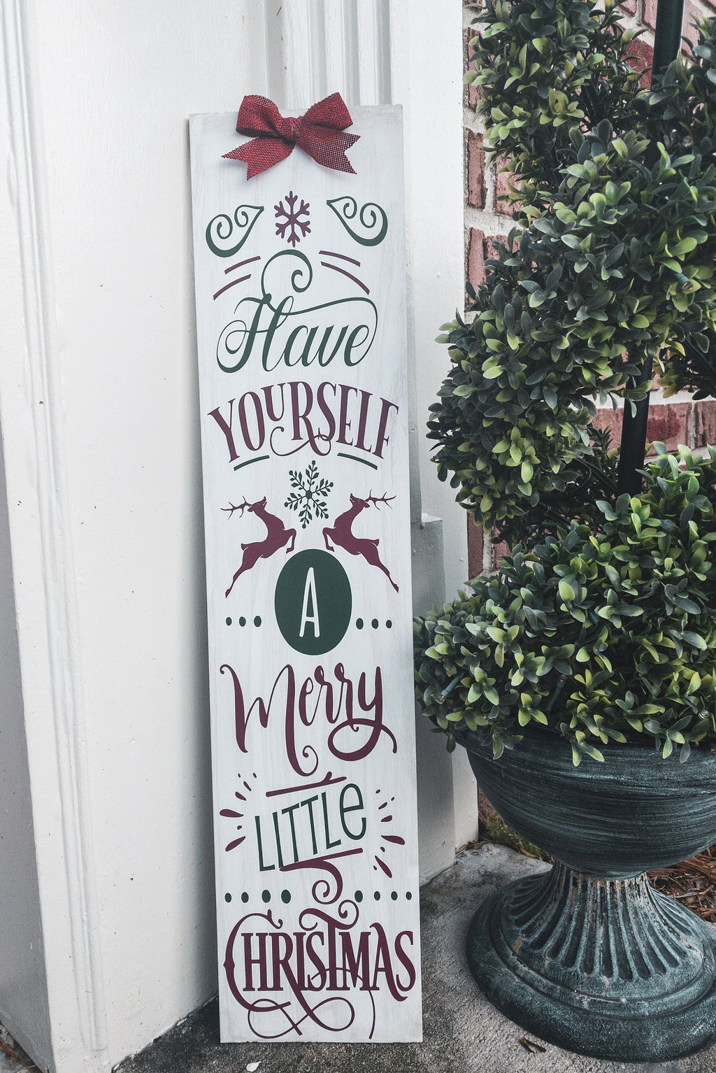 Have Yourself a Merry Little Christmas rustic wood porch leaner sign with red burlap bow on white wood wall beside Christmas garland with ornaments
