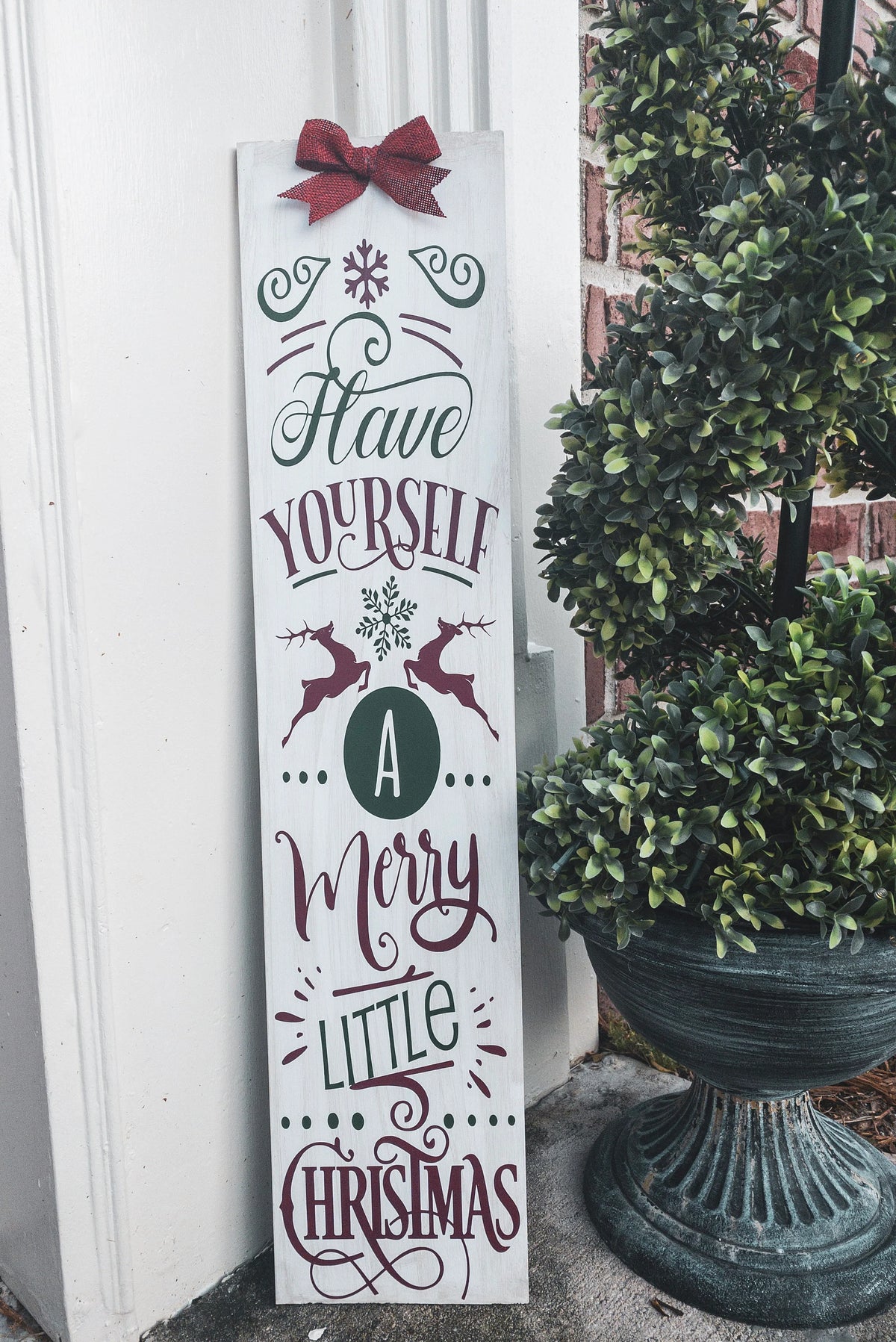 Have Yourself a Merry Little Christmas rustic wood porch leaner sign with red burlap bow on white wood wall beside Christmas garland with ornaments