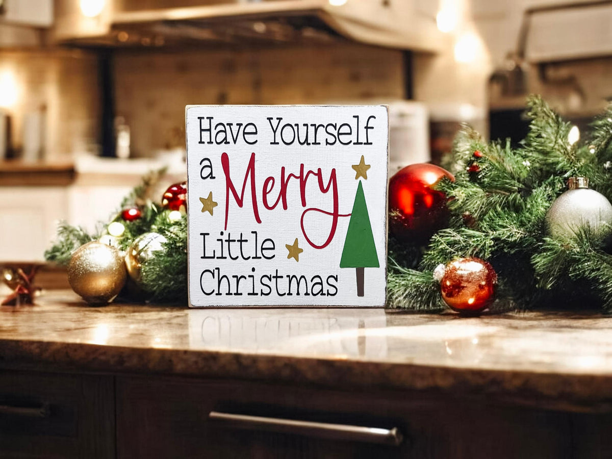 Have Yourself a Merry Little Christmas white wood block sign on wood counter with red and gold ornaments and Christmas tree bokeh