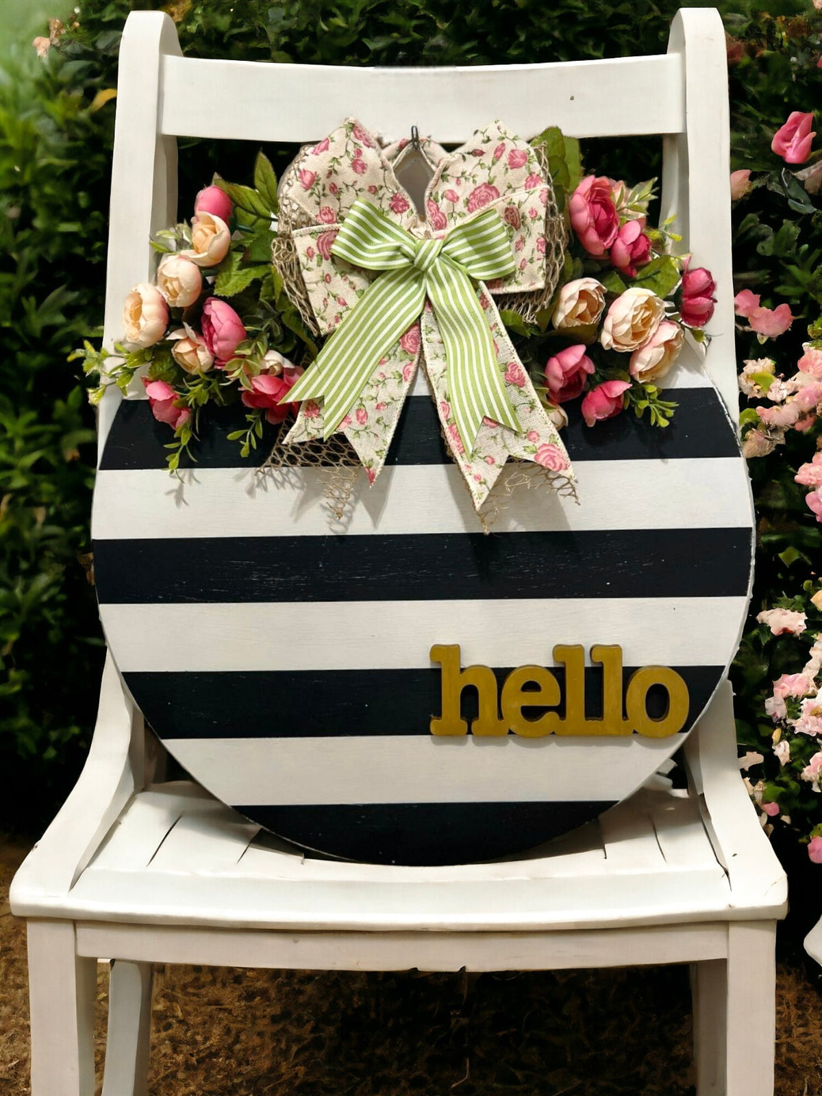 Hello black and white striped round door hanger with pink floral bow on black front door of white farmhouse with potted mums