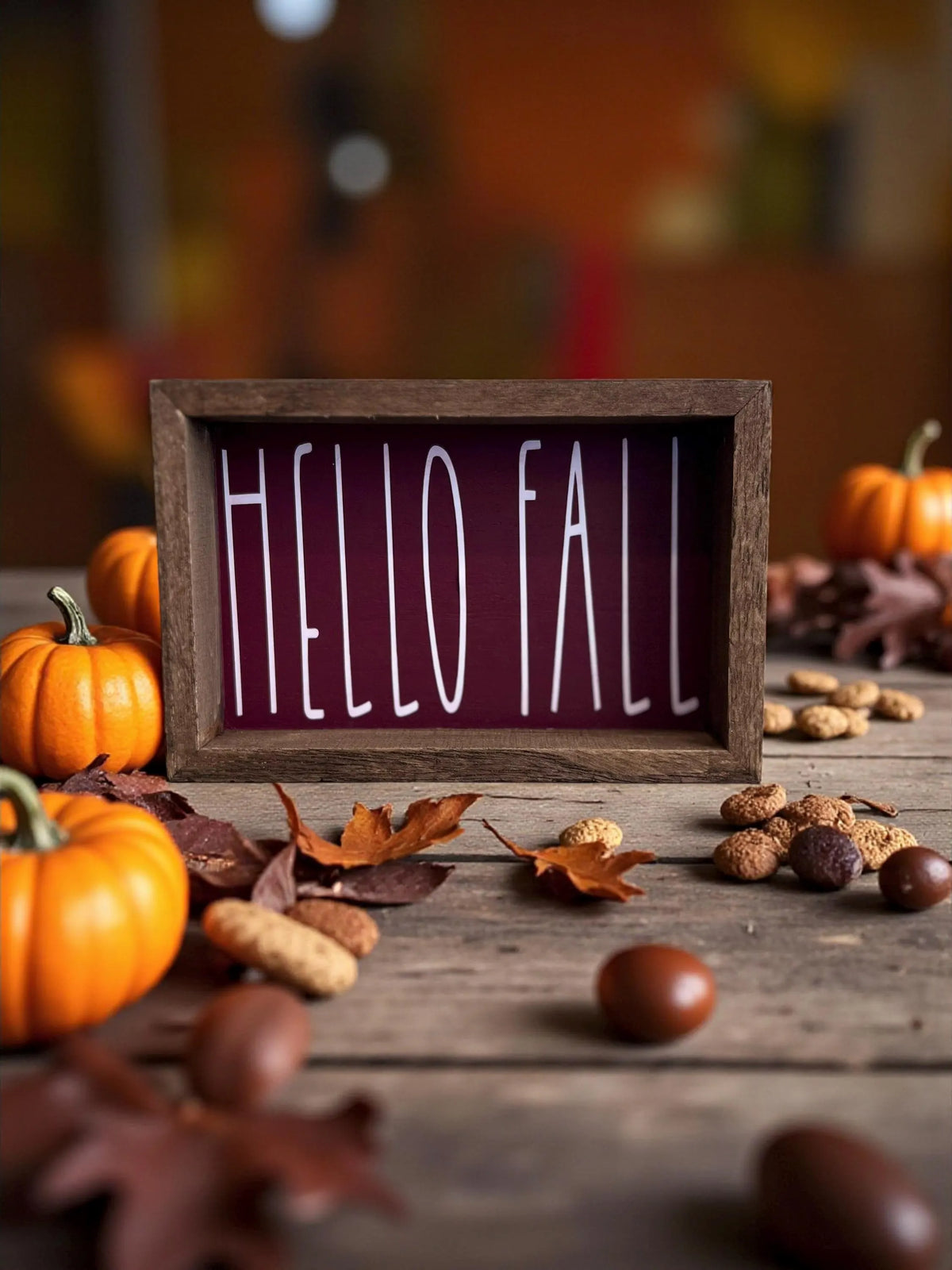Hello Fall cranberry framed wood sign on dark wood table surrounded by orange pumpkins pine cones and fall leaves