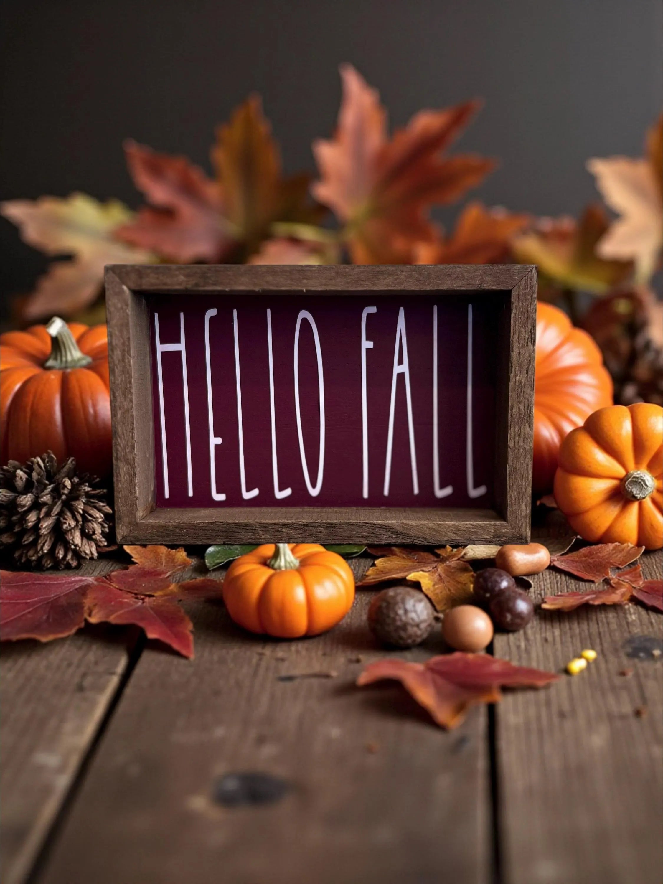 Hello Fall cranberry framed wood sign on rustic wood table surrounded by mini pumpkins autumn leaves and acorns