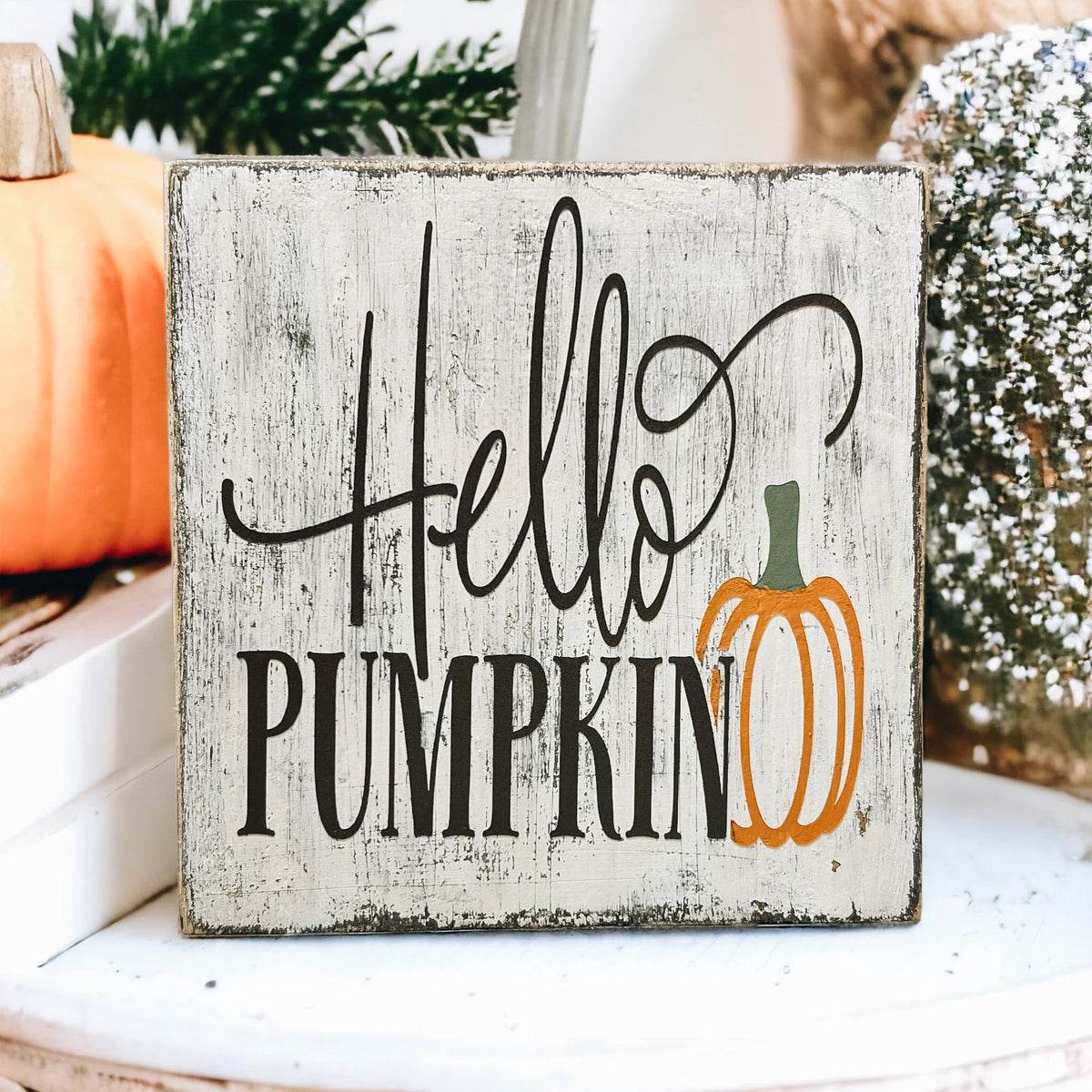 Hello Pumpkin wood block sign with black script and serif lettering and orange pumpkin graphic on distressed white pine on white wood surface with orange pumpkins and green leaves porch hero shot