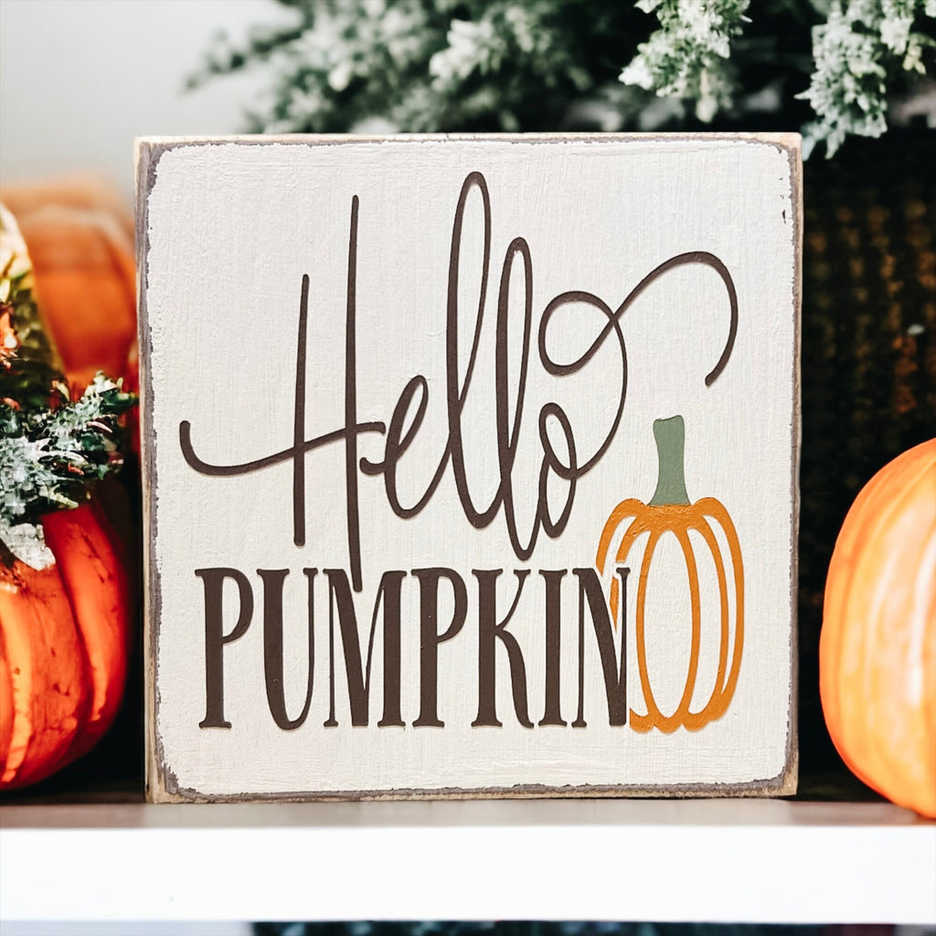Hello Pumpkin wood block sign with black script and serif lettering and orange pumpkin graphic on distressed white pine on white cake stand with orange pumpkins and pine cones angled view
