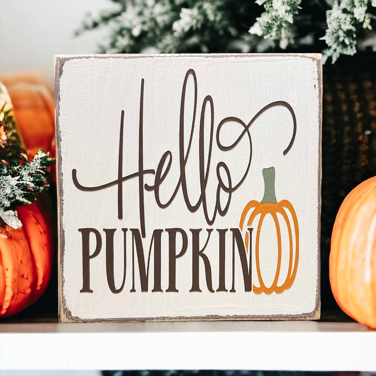 Hello Pumpkin wood block sign with black script and serif lettering and orange pumpkin graphic on distressed white pine on white cake stand with orange pumpkins and pine cones angled view