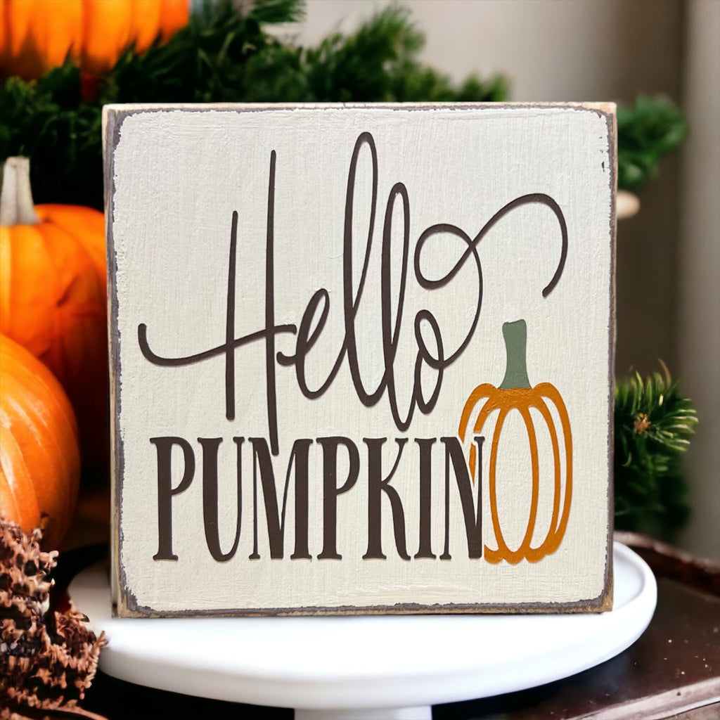 Hello Pumpkin wood block sign with black script and serif lettering and orange pumpkin graphic on distressed white pine on white cake stand with white and orange pumpkins and pine cones close up