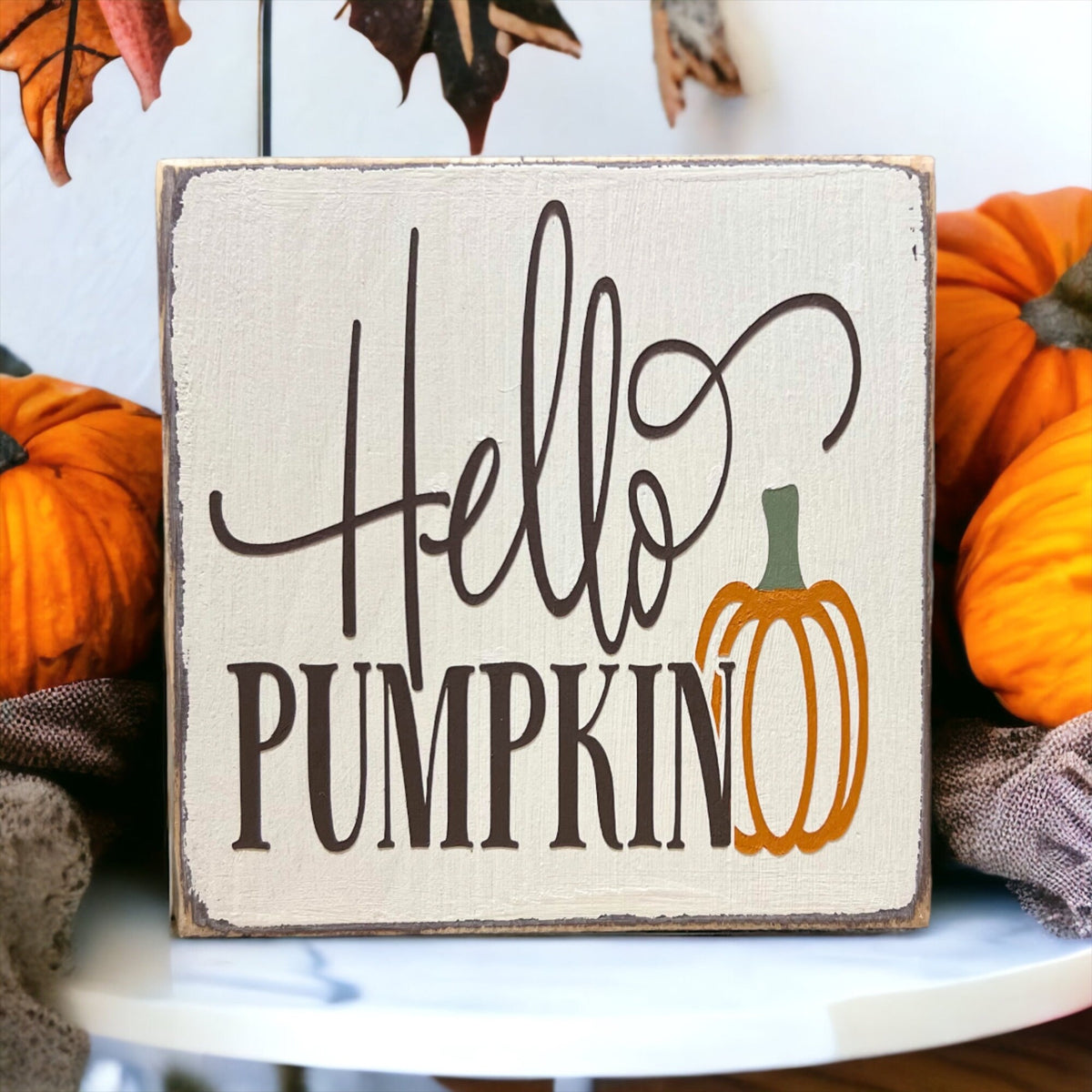 Hello Pumpkin wood block sign with black script and serif lettering and orange pumpkin graphic on distressed white pine on white cake stand with orange pumpkins and pine cones rustic wood shelf