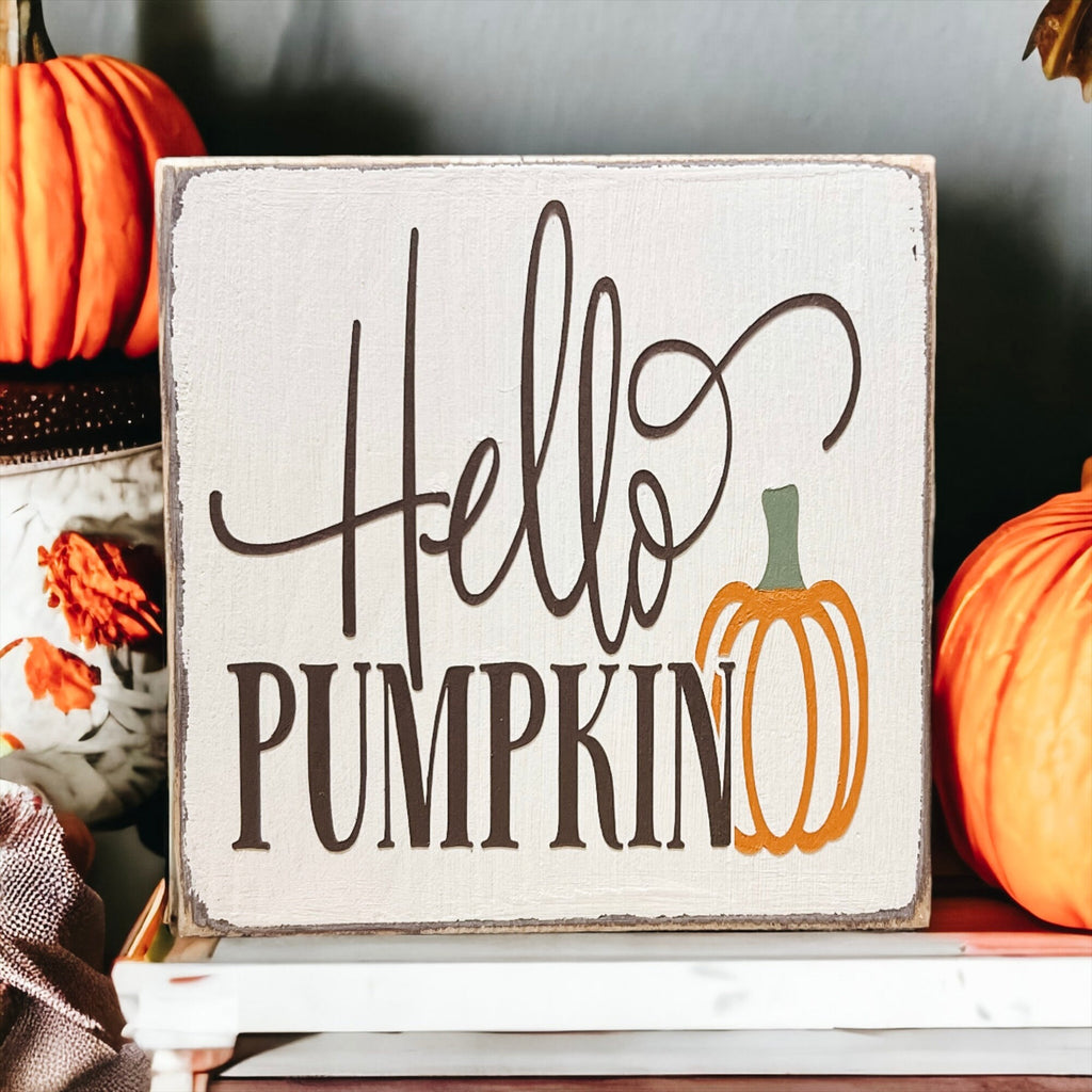 Hello Pumpkin wood block sign with black script and serif lettering and orange pumpkin graphic on distressed white pine on white mantel shelf with orange and black pumpkins and dried botanicals