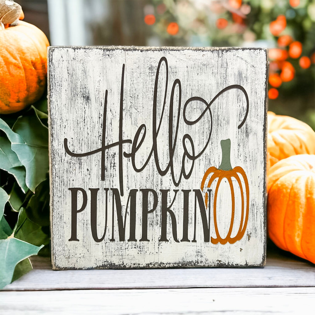 Hello Pumpkin wood block sign with black script and serif lettering and orange pumpkin graphic on distressed white pine on white cake stand with orange pumpkins and frosted pine greenery