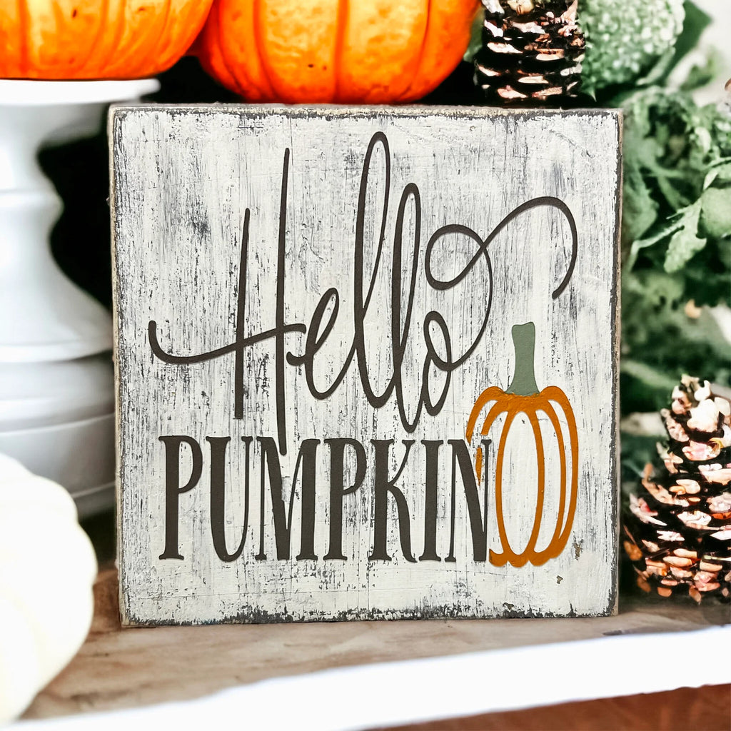 Hello Pumpkin wood block sign with black script and serif lettering and orange pumpkin graphic on distressed white pine on white cake stand with orange pumpkins and pine greenery