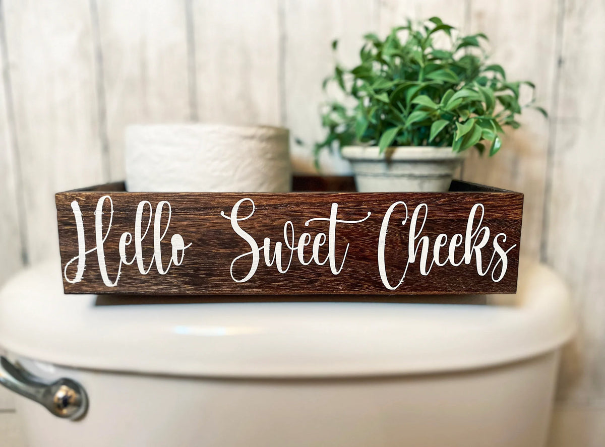 Hello Sweet Cheeks dark stained farmhouse toilet paper box on toilet tank with plant and toilet paper roll close up