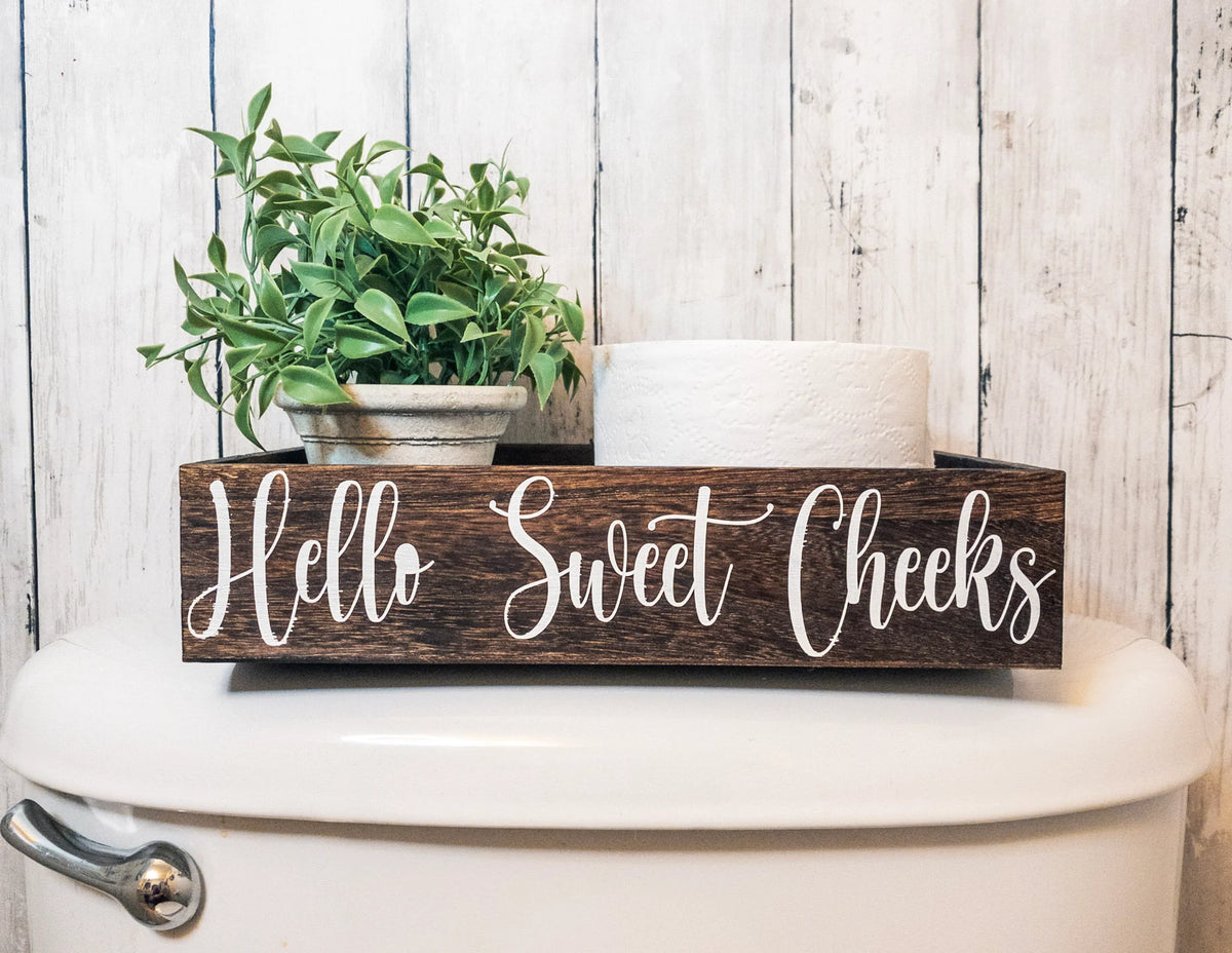 Hello Sweet Cheeks dark stained farmhouse toilet paper box on toilet tank with plant and toilet paper roll wide view