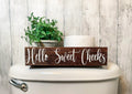 Hello Sweet Cheeks dark stained farmhouse toilet paper box on toilet tank with plant and toilet paper roll front view