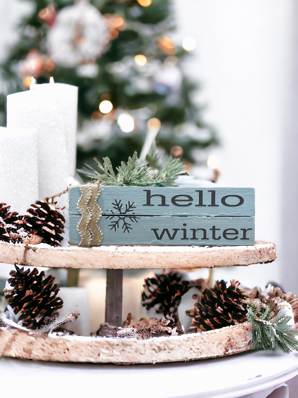 Hello Winter dusty blue wood book stack with snowflake and burlap ribbon on tiered tray with pine cones and white candles