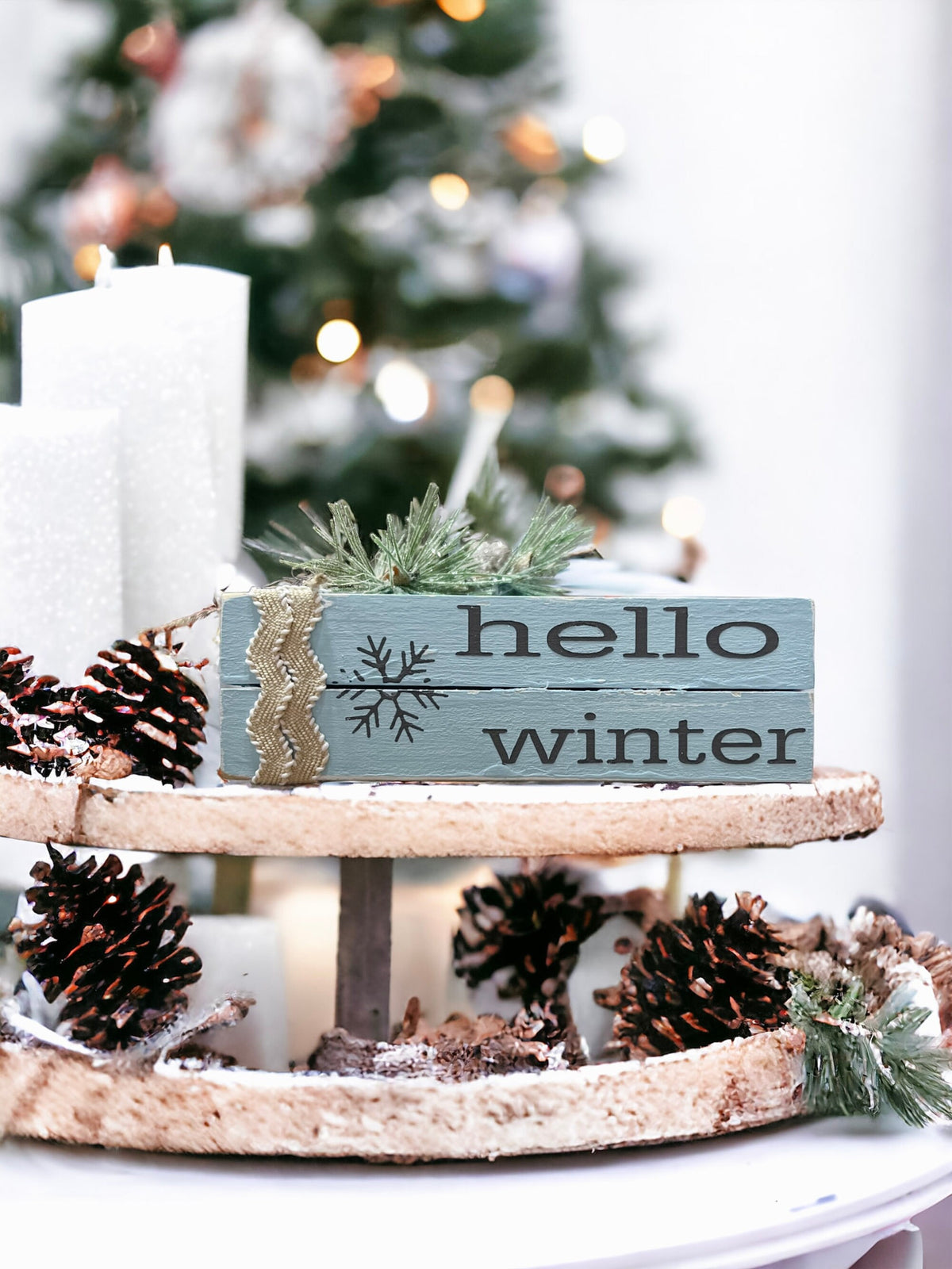 Hello Winter dusty blue wood book stack with snowflake and burlap ribbon on tiered tray with pine cones and white candles