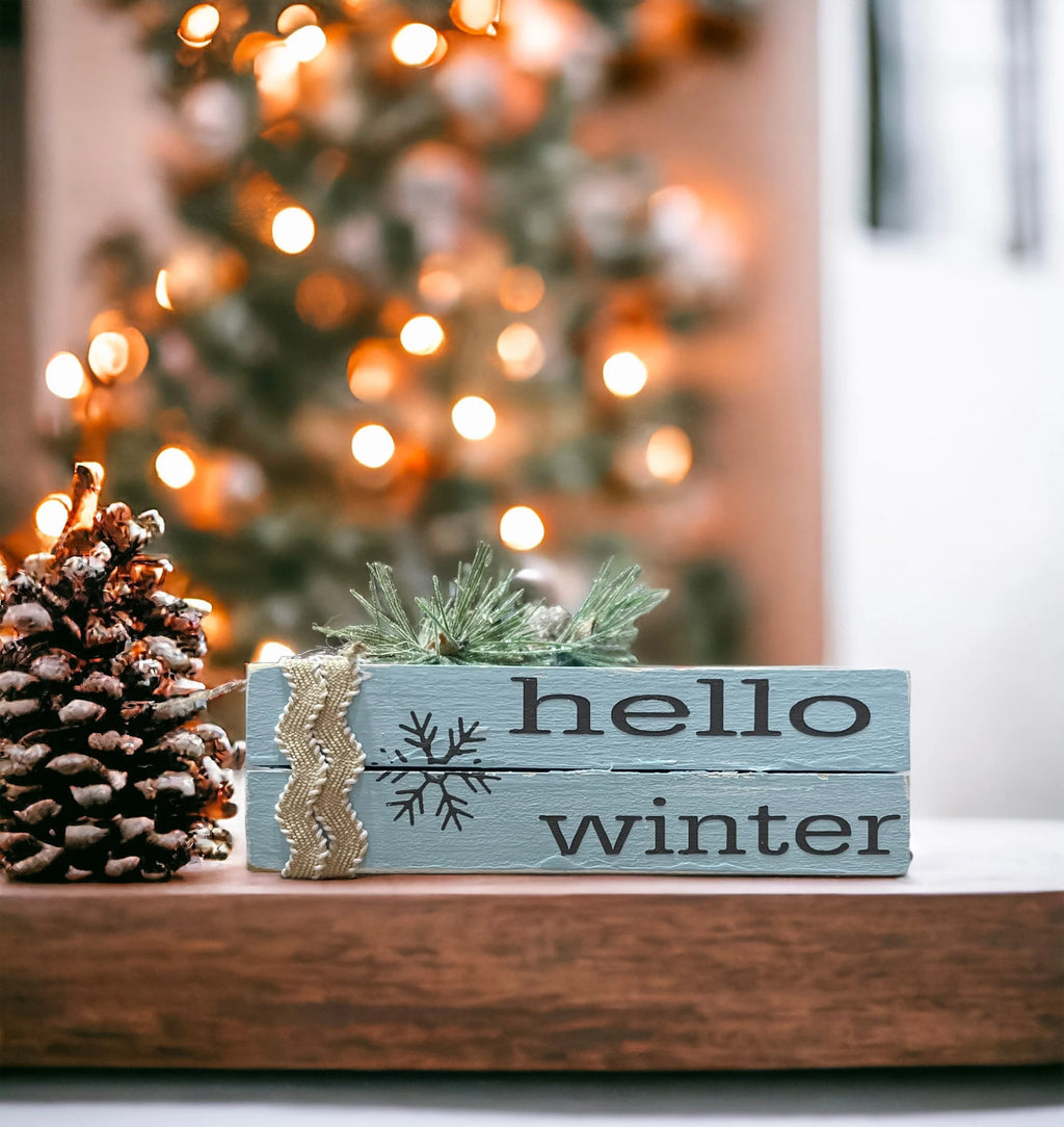 Hello Winter dusty blue wood book stack with snowflake and burlap ribbon on white tiered tray with pine cones and Christmas ornaments