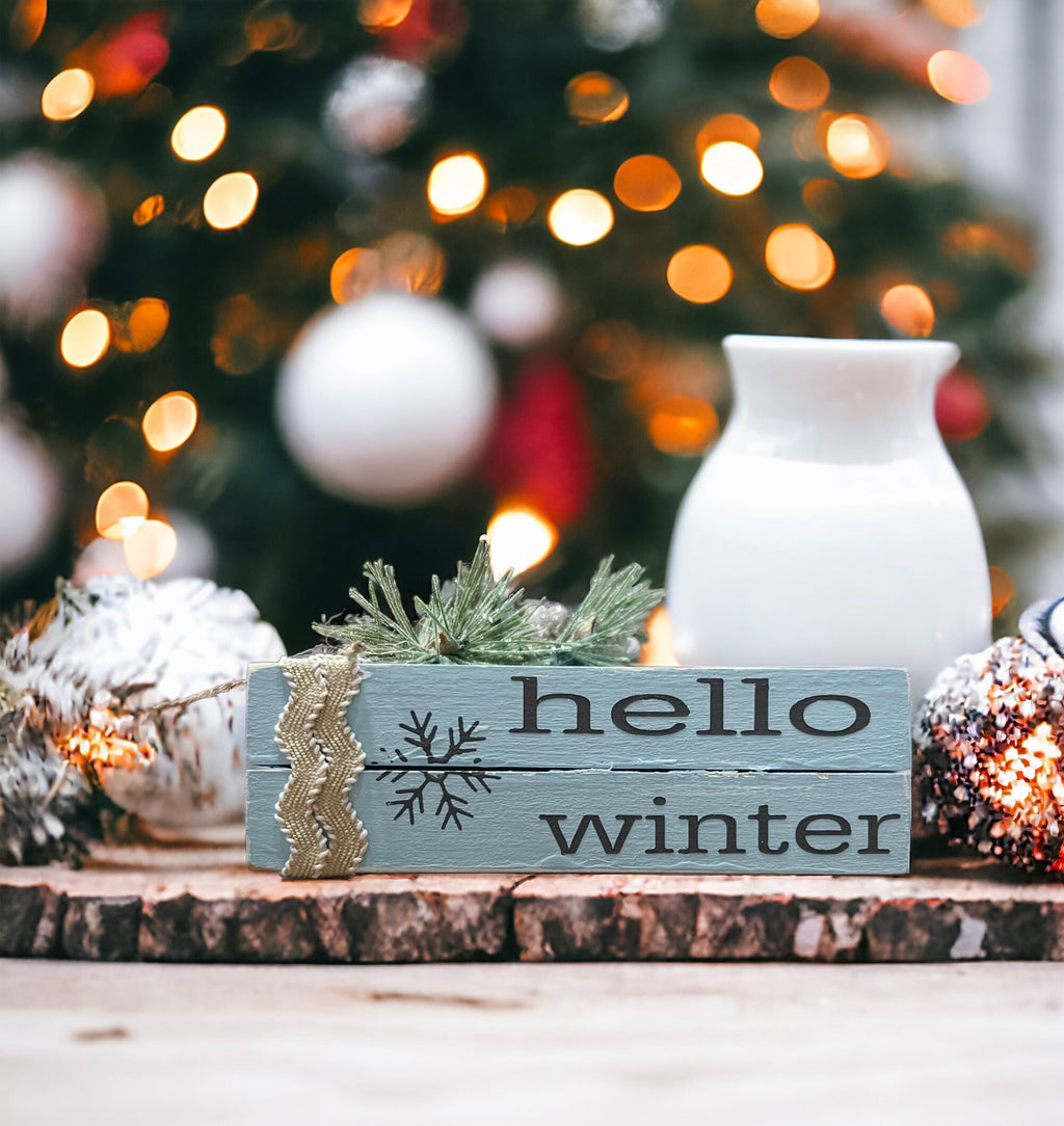 Hello Winter dusty blue wood book stack with snowflake and burlap ribbon on wood shelf with pine cone and Christmas tree bokeh