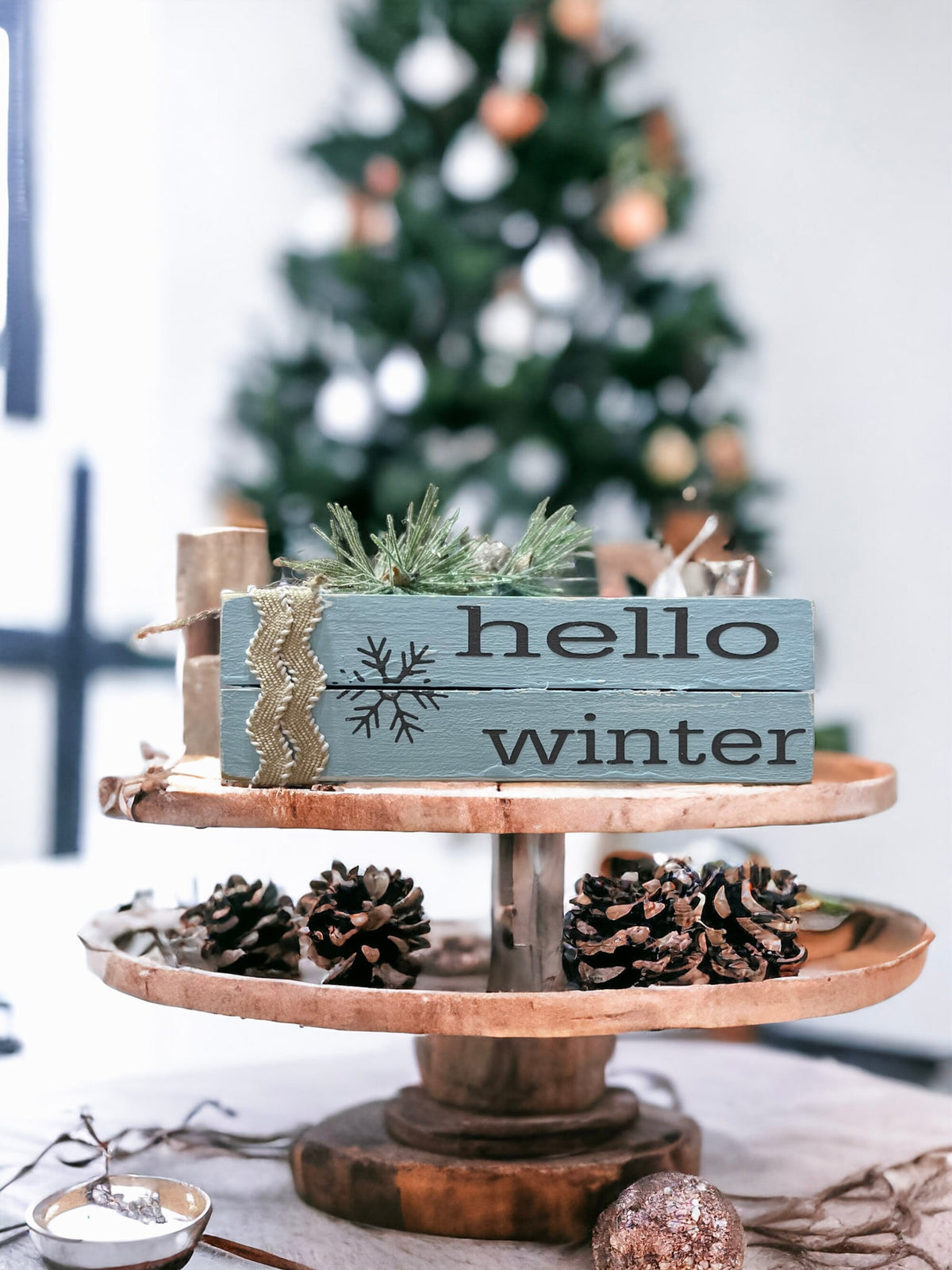 Hello Winter dusty blue wood book stack with snowflake and burlap ribbon on wood slice with Christmas tree bokeh lights