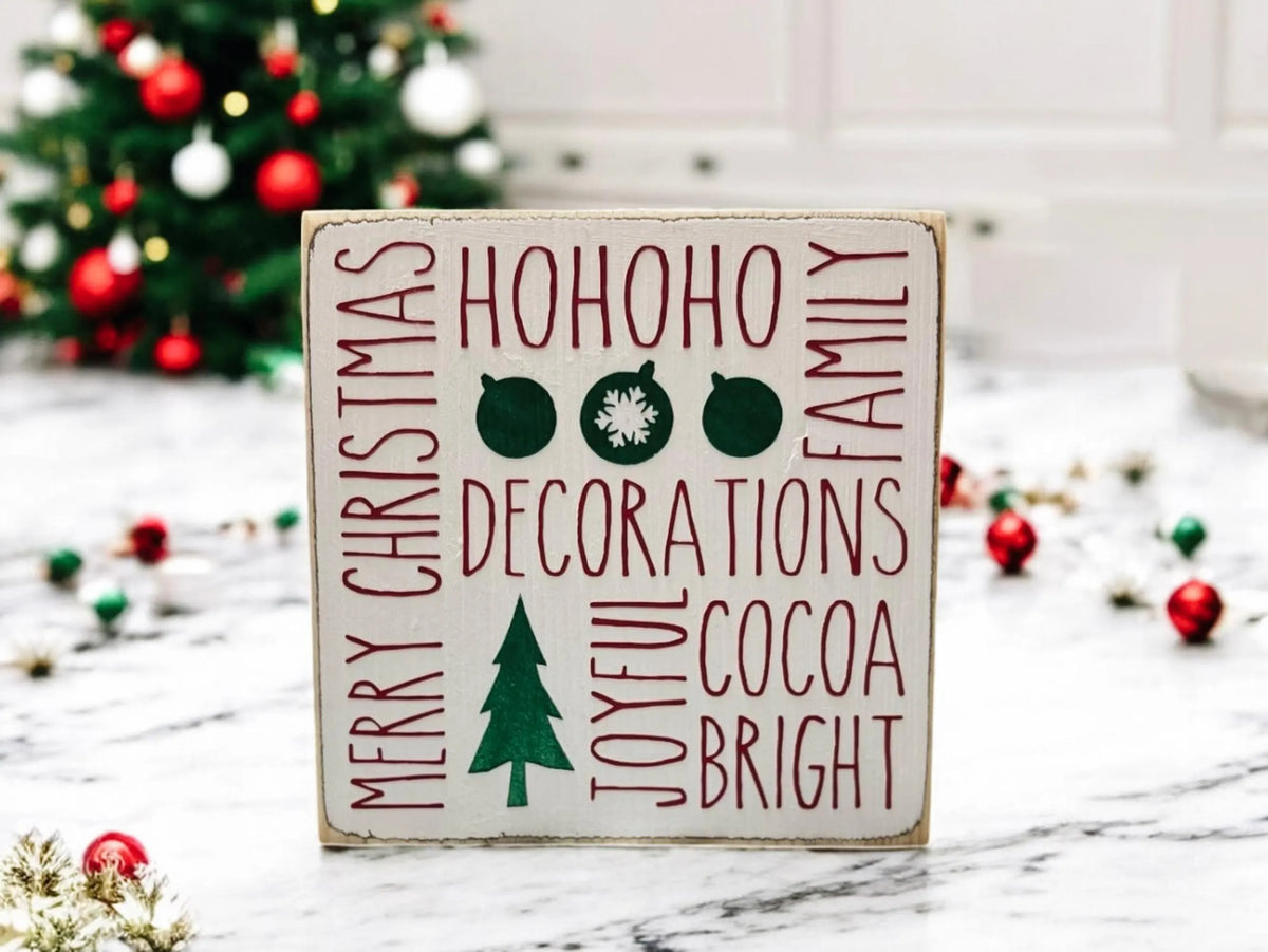 Christmas sayings wood block sign with HoHoHo Merry Christmas Family Decorations Joyful Cocoa Bright in red and green on distressed white pine on white surface with pine cones and string lights side angle