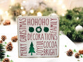 Christmas sayings wood block sign with HoHoHo Merry Christmas Family Decorations Joyful Cocoa Bright in red and green on distressed white pine on white surface with pine cones and string lights wide shot
