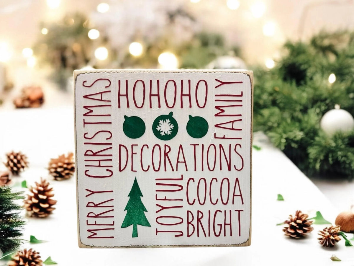 Christmas sayings wood block sign with HoHoHo Merry Christmas Family Decorations Joyful Cocoa Bright in red and green on distressed white pine on white surface with pine cones and string lights wide shot