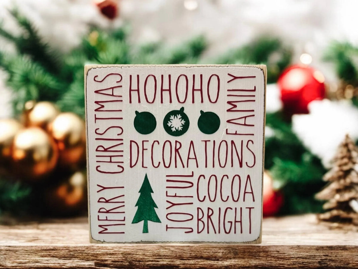 Christmas sayings wood block sign with HoHoHo Merry Christmas Family Decorations Joyful Cocoa Bright in red and green on distressed white pine on rustic wood surface with lit Christmas tree bokeh close up